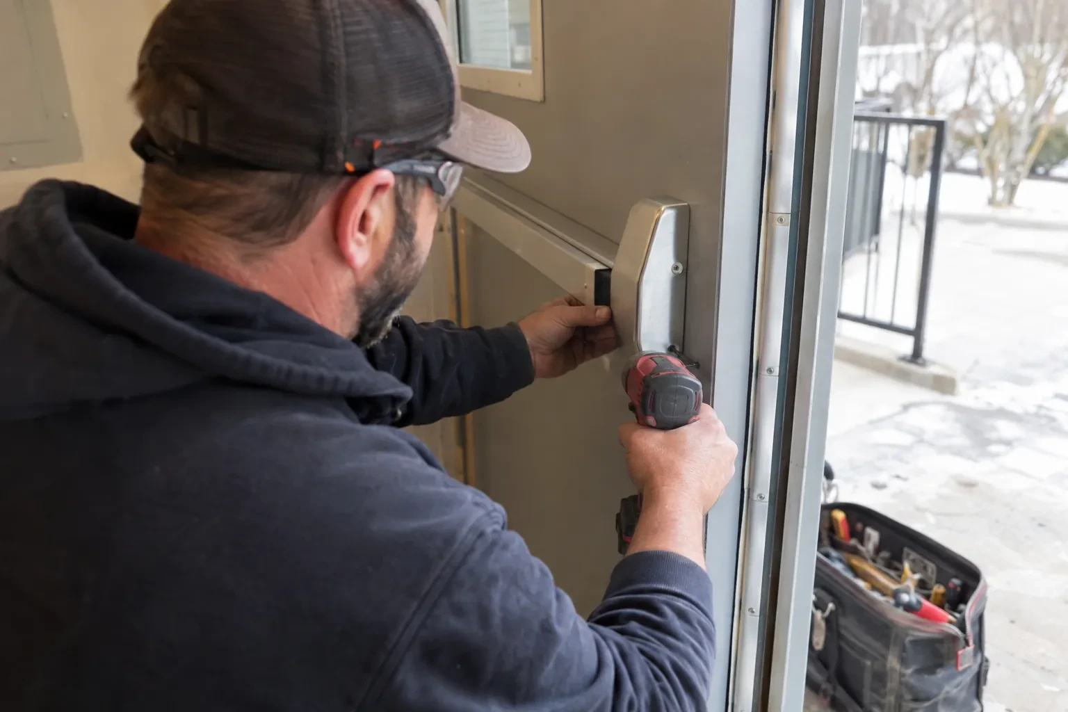Technician in a cap and safety glasses holding a push bar end cap against a metal door with a red drill.